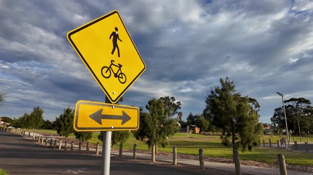 Slow motion view of a warning sign with pedestrian and bicycle symbols above a double-headed arrow plate, where a shared path meets or crosses a road, to warn drivers to slow down and look both ways. 