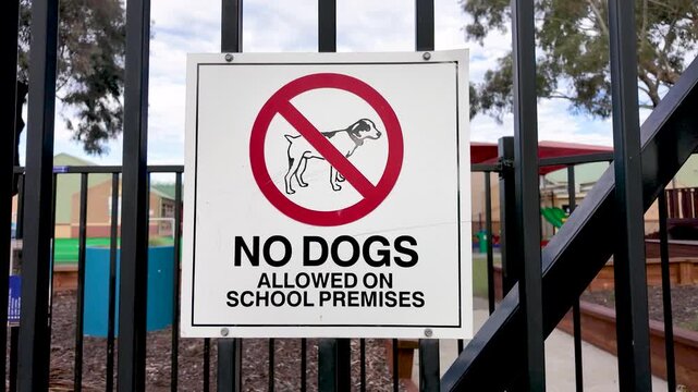 a "No Dogs Allowed on School Premises" sign with a red prohibition symbol, on a security fence at a primary school entrance, school buildings blurred behind. Pet and animal restrictions in Australia.
