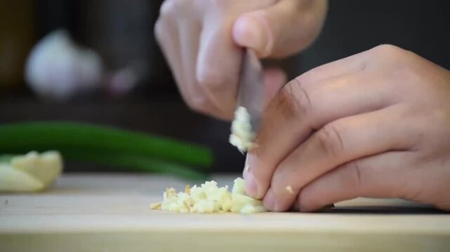 Close-up of hands mincing garlic on a wooden cutting board with a chef's knife in a kitchen setting