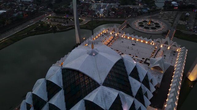 Aerial view of the illuminated Al Jabbar Grand Mosque with its unique geometric dome and surrounding reflecting pool at dusk in Bandung, West Java, Indonesia.