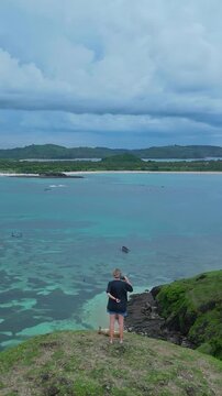 Caucasian woman cliff watching stormy bay dog resting beside, overcast sky and heavy clouds, green coastal slopes and rocky cove, smartphone held up to frame distant horizon, Lombok island