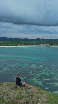 Woman sits with dog watching storm over turquoise bay, heavy clouds gather on horizon above calm water, introspective mood, Bukit Merese, Lombok, Tanjung Ann, vertical video