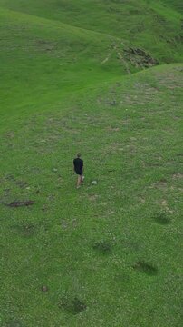 Woman walking on green hill, solitary hiker crosses lush grassy slope in aerial view, slow steady pace, black jacket and short hair, scattered stones and faint footpath, wide panoramic