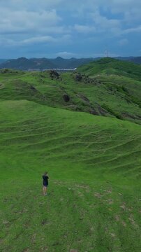 White woman walking hilltop overlooking bay solo photographer surveys sweeping green valleys and hidden sandy cove dramatic ridgelines aerial perspective adventurous tone, Bukit Merese, Lombok