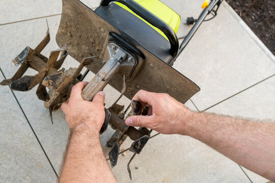 A person is adjusting the tines of a garden tiller on a patio surface. Hands are seen modifying the attachment of a tiller used for cultivating soil in an outdoor setting.