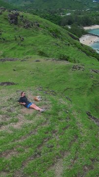 Woman and dog on cliff overlooking turquoise bay, reclining on green grass with panoramic ocean view, sandy beach below, Bukit Merese, Lombok, Tanjung Ann, vertical video