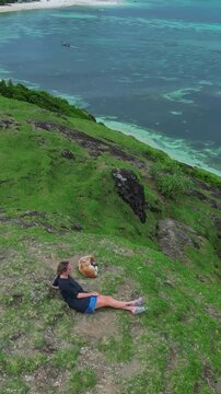 Woman sits with dog watching storm over turquoise bay, heavy clouds gather on horizon above calm water, introspective mood, Bukit Merese, Lombok, Tanjung Ann, vertical video