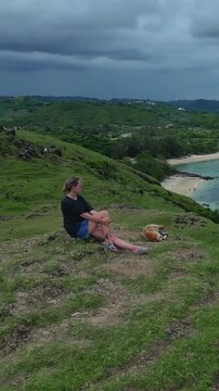 Woman sits with dog watching storm over turquoise bay, heavy clouds gather on horizon above calm water, introspective mood, Bukit Merese, Lombok, Tanjung Ann, vertical video