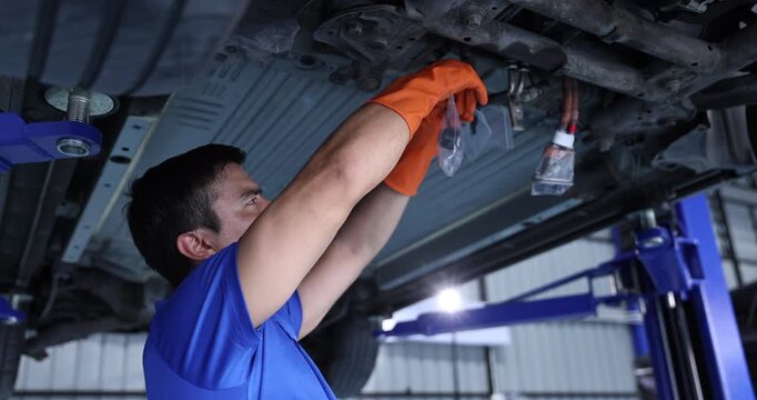 Automotive technician wearing orange gloves inspects battery connections beneath a lifted electric vehicle for residual moisture after flooding. 