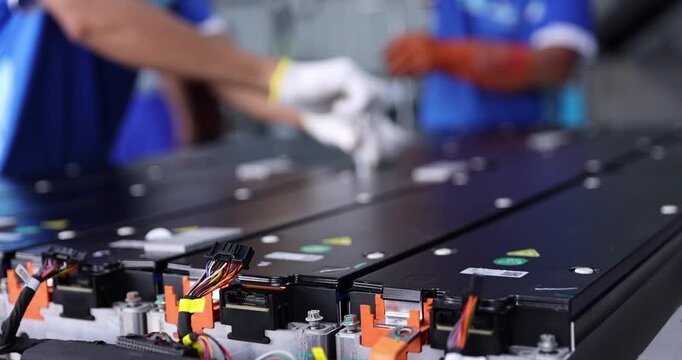 Two technicians in blue workwear use hand tools to tighten fasteners on electric vehicle battery modules. Close-up of gloved hands working together during battery pack assembly .