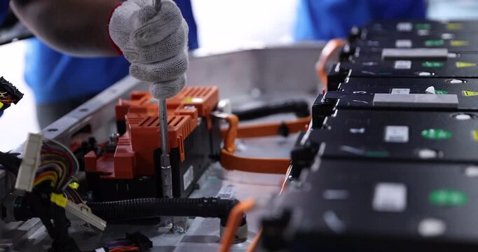 Close-up of a gloved technician using a hand tool to tighten fasteners inside an electric vehicle battery pack. Battery modules, orange electrical components.
