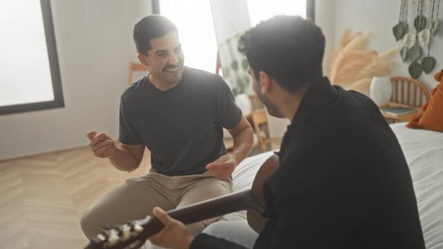 Man sitting on bed with acoustic guitar, smiling and tapping his hand while a friend plays guitar in a building; warm friendship music.
