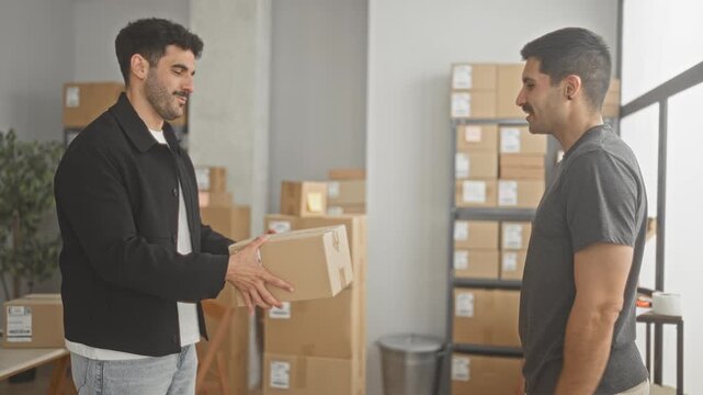 Two hispanic men hand a shipping box to each other and later shake hands while smiling among stacked parcels and shelving in a small business storage building; teamwork trust.
