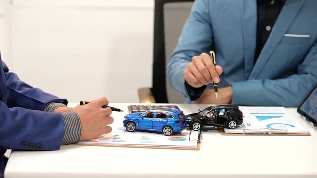 Car dealership agents discuss paperwork beside blue and black model cars, showing vehicle purchase, insurance, financing, or sales agreement.