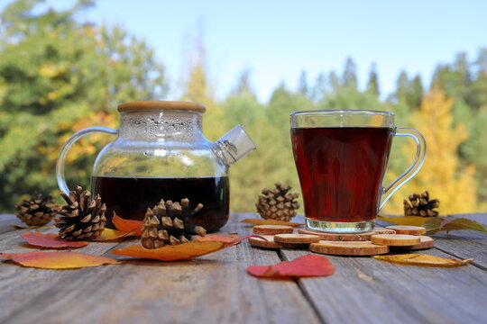 On a wooden table there are a glass mug of hot black tea and a glass teapot amidst foliage and pine cones, surrounded by an autumn forest landscape. 