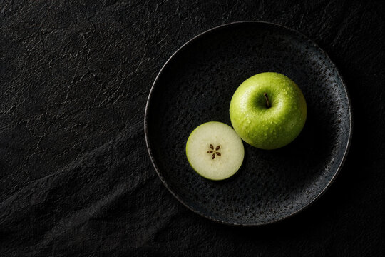 A green apple and a sliced apple rest on a dark ceramic plate over a black textured surface, creating a minimalist food composition.