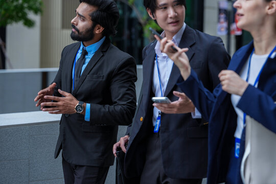 Three colleagues walking in profile point ahead while explaining an idea on the move under fresh trees in the public realm of a smart city district, a pale stone parapet runs behind them