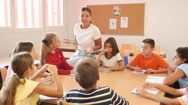 Friendly smiling teacher explaining studying material to diligent tweens sitting at common table and writing in notebooks during lesson at school 