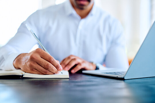 Man's hands writing notes in a small book on a desk next to a laptop, representing work, organization, and ideas
