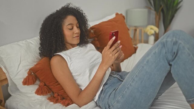 Woman lying on bed in bedroom holding red smartphone and scrolling with pursed lips, orange tassel pillow and lamp visible; relaxed leisure.