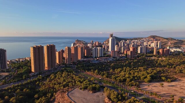 The skyline of Benidorm set against the ocean as the sun sets. The area features buildings and greenery along the coastline, highlighting the beauty of the region.