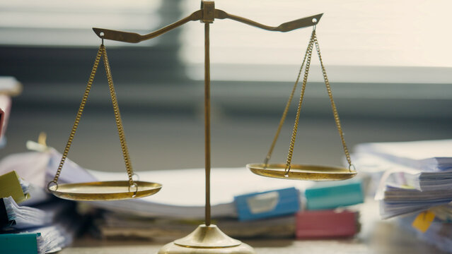 Golden scales sit atop stacks of papers and folders. Soft focus blurs the background, drawing attention to the balance. The symbol of justice rests in a cluttered office space. No people are present