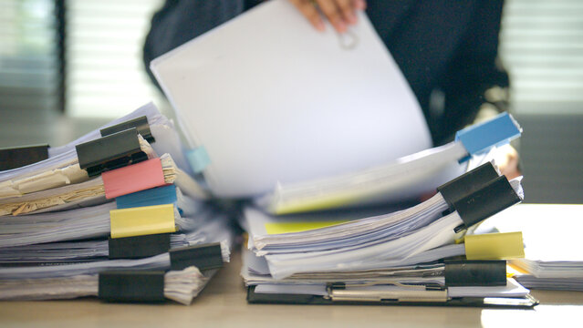 Busy office scene with towering stacks of papers. Person holds documents, suggesting work pressure or deadline stress. Colorful tabs add visual contrast to the cluttered desk