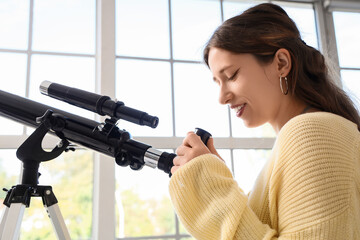 Fototapeta premium Stem education and scientific discovery, young woman looking through a telescope to explore astronomy and space science in a bright home interior