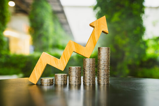 Economic growth and upward financial trend concept, large wooden zigzag arrow pointing up beside ascending coin stacks on table, symbolizing revenue acceleration, profit, and investment.