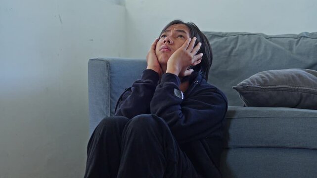 Young man holding his head and gazing upward in deep worry and distress sits on floor by sofa for bipolar disorder concept