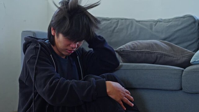 Young man weeping and burying face in hands reflecting bipolar depressive agony sits on floor by sofa