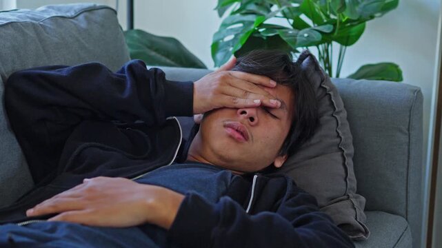 Young man lies on sofa with hand covering eyes portraying severe physical and mental exhaustion from bipolar depression