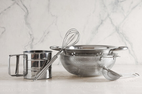 Home cooking and baking preparation stainless steel kitchen tools including flour sifter whisk colander and strainer on a marble surface