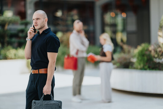 A businessman stands outdoors talking on his smartphone while holding a briefcase. Two blurred colleagues speak in the background, suggesting a professional urban setting.