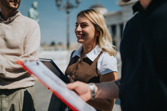 A smiling young woman holds a folder and listens to colleagues during an outdoor conversation. The small group stands by a city landmark discussing documents and ideas.