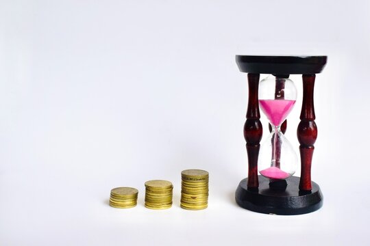 Incremental stacks of gold coins alongside a vintage wooden hourglass with pink sand over a plain white background.