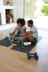 Fototapeta premium African American couple sitting on mats in living room in workout gear holding phone and weights