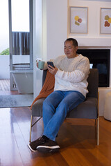 Fototapeta premium Senior Chinese man sitting at home on gray chair holding smartphone and mug, bathtub visible