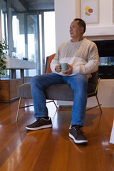 Fototapeta premium Senior Asian man sitting on grey chair at home wearing cream sweater holding light green mug