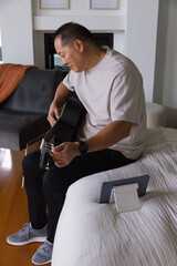 Fototapeta premium Senior Asian man sitting on bed edge in bedroom living area holding acoustic guitar, tablet nearby