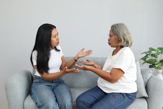 A young woman and a senior Asian woman sitting on a sofa at home, having an intense conversation, discussing a problem, or having an argument with expressive hand gestures.