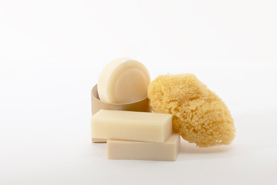 Minimalist spa collection featuring artisanal cream-colored soap bars and a round solid cleanser. A natural sea sponge adds organic texture against the clean white background. Ideal for eco-luxury.