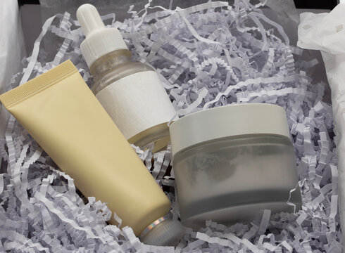 Abstract skincare gift set featuring a facial cream jar, serum dropper bottle, and a soft yellow tube nestled in white crinkled paper shreds.
