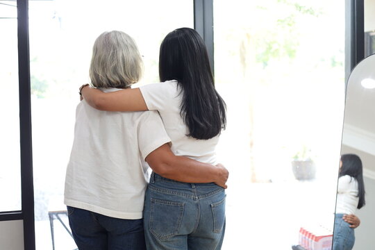 A rear view of a young woman and a senior Asian woman standing together, embracing each other with arms around shoulders, showcasing love, care, and family bonding at home.
