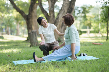 Fototapeta premium Senior Asian woman practicing assisted stretching on a yoga mat with a younger woman in a park, representing rehabilitation, flexibility, healthy aging, and wellness.