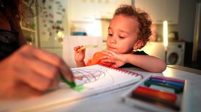 Cute toddler with curly hair concentrating on drawing with colorful crayons in his sketchbook with his mother, creating artwork for the halloween holidays in a cozy kitchen atmosphere at night