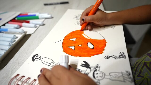 Young girl's hands coloring a spooky jack o' lantern drawing on a sketchbook with an orange felt tip pen, getting ready for the halloween celebration with creative artistic activities at home
