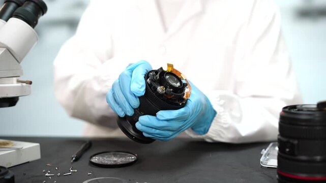 Technician in blue gloves repairs a camera lens beside a microscope, handling exposed lens parts on a workbench in a clean laboratory