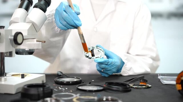 Forensic scientist in blue gloves uses a brush to collect fingerprint evidence beside a microscope in a crime laboratory.