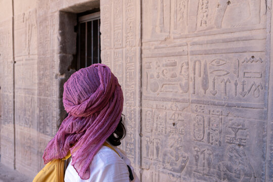 Woman Solo traveler wearing the Egyptian turban or Imma, observing hieroglyphs and historical reliefs inside an ancient egyptian temple, immersing in cultural heritage